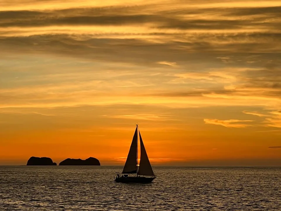 A sailboat under sail at sunset off Playa Potrero, silhouetted against a glowing orange sky with the Catalina Islands in the distance, Guanacaste, Costa Rica