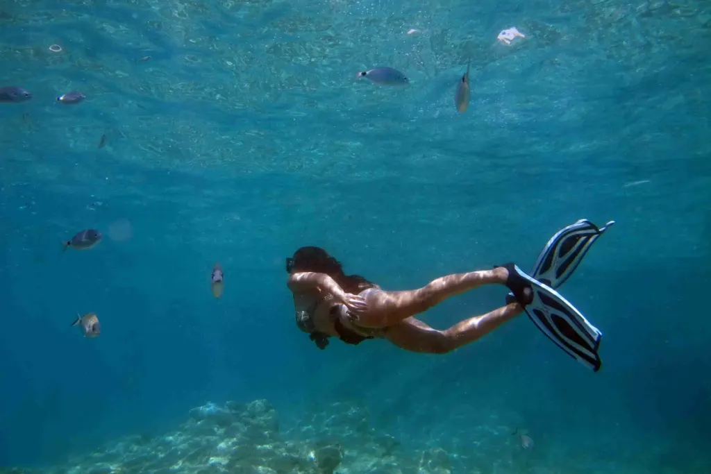 A woman snorkeling in a swimsuit and fins glides over a rocky reef surrounded by tropical fish in the clear turquoise water off Playa Potrero, Costa Rica