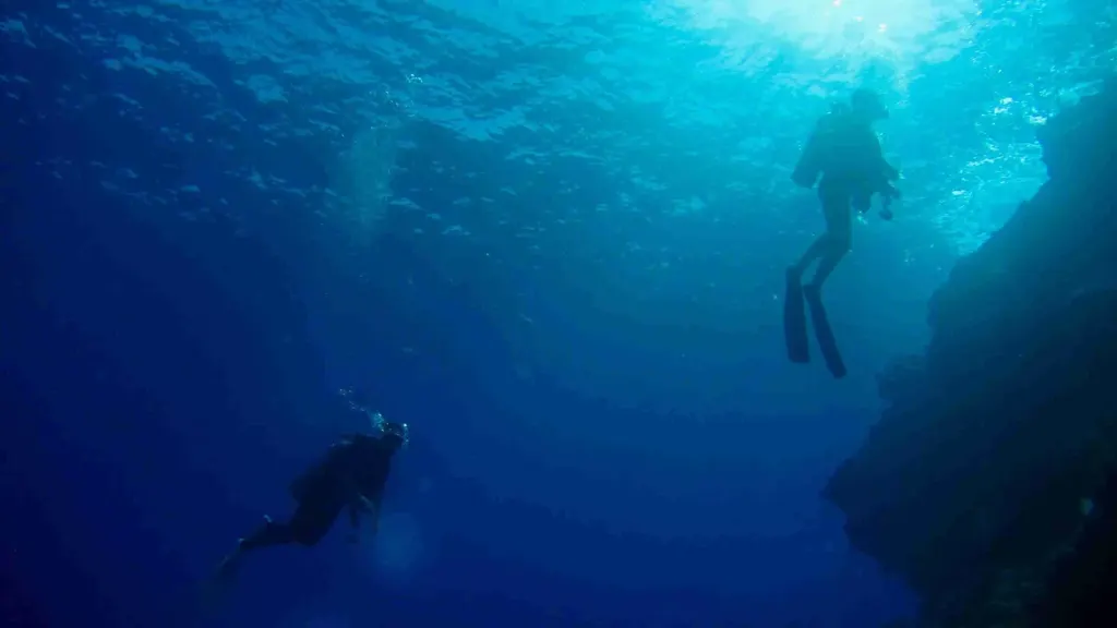 Two scuba divers descending along a deep volcanic reef wall toward the sunlit surface, scuba diving off Playa Potrero, Costa Rica