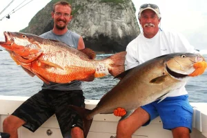 Two anglers holding a large cubera snapper and an amberjack caught with Fish Potrero, with a rocky island behind off the Guanacaste coast, Costa Rica
