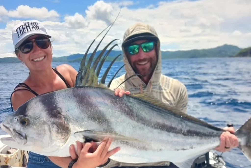 A couple holding a large roosterfish with its distinctive comb-like dorsal fin fanned up, caught inshore with the Guanacaste coastline behind, Costa Rica